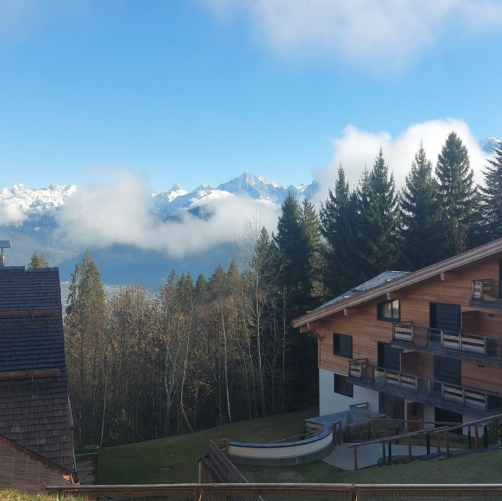 Un bâtiment moderne à l’extérieur en bois, doté d’un balcon, et entouré d’une forêt, avec des montagnes en arrière-plan.