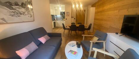 A modern living room with a sofa, armchairs, a coffee table, and a TV. There is a dining area with a table and chairs in the background.