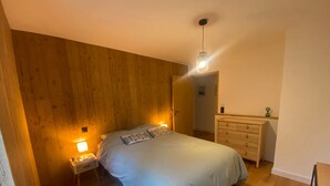 A bedroom with a wooden wall, a bed with pillows, a nightstand, a dresser, and a hanging light fixture.