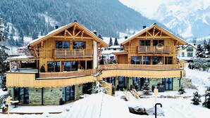 Un chalet en bois doté d’un balcon et d’une base en pierre, entouré d’arbres et de montagnes couverts de neige.