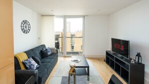Un salon moderne avec un canapé noir, une table basse, un téléviseur posé sur un meuble noir et une grande fenêtre donnant sur des immeubles.
