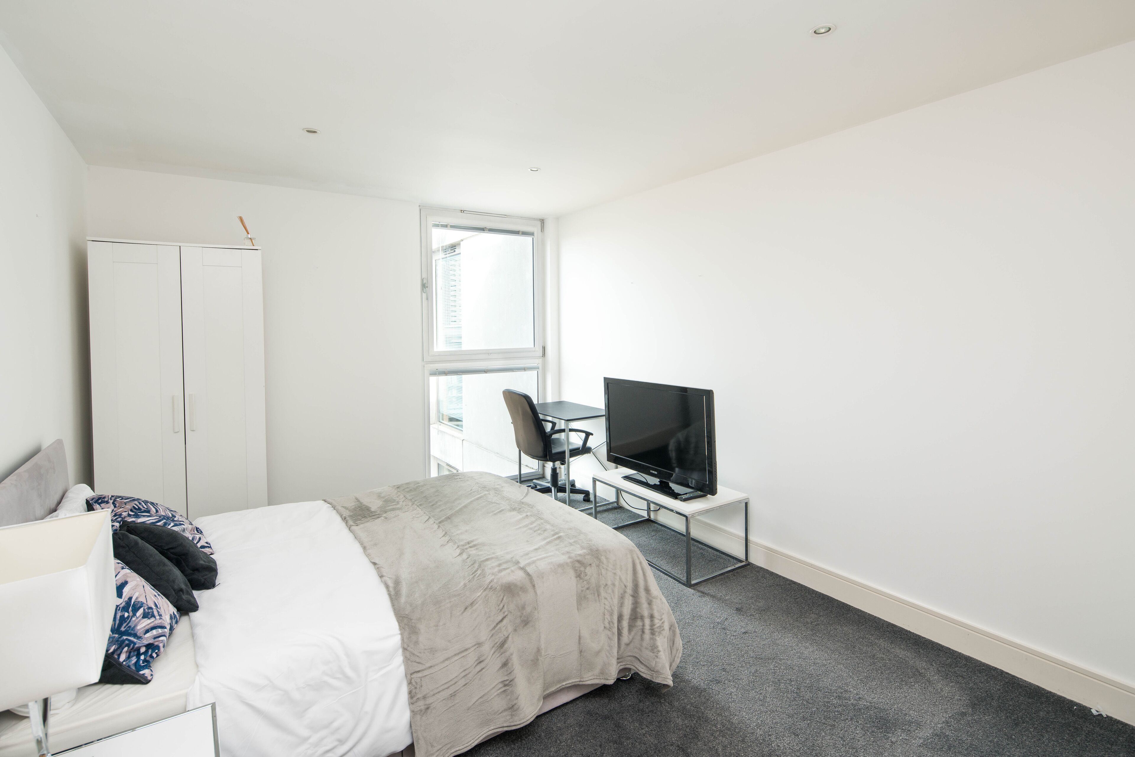A bedroom with a bed, a desk with a TV, a wardrobe, and a window.