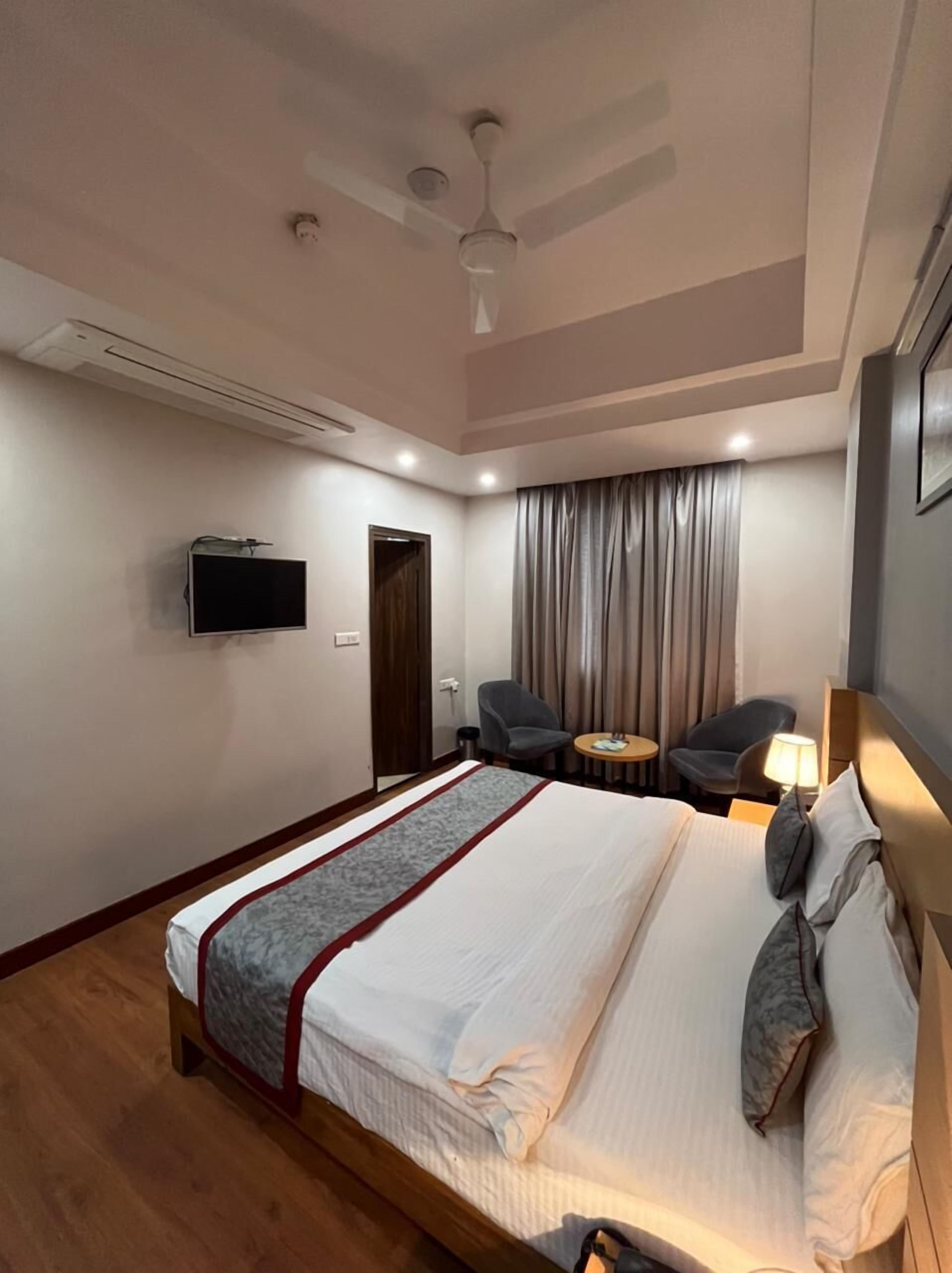 A hotel room with a bed, a TV, a ceiling fan, a desk with a chair, and a window with curtains.