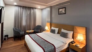 A hotel room with a large bed, two bedside tables, a ceiling fan, a desk with a chair, and a window with curtains. - Hotel Sarvin (Lucknow)
