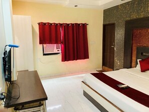 A hotel room with a bed, a desk with a computer, a window with curtains, and a wall-mounted TV. - Hotel Jagdish By GRB (Raipur)