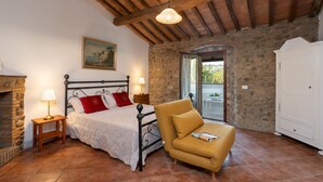 A bedroom with a stone wall, a bed with red pillows, a wooden chair, a nightstand with a lamp, and a painting on the wall. - Il Pratone 12 Emma Villas (Pomarance)