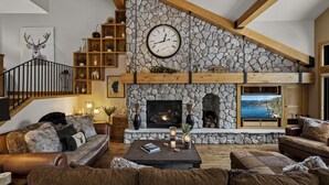 A cozy living room with a stone fireplace, a large clock, and a television displaying a scenic view.