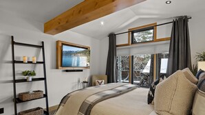 A bedroom with a large bed, a wooden ceiling beam, a wall-mounted TV, and a window with a view of a lake.