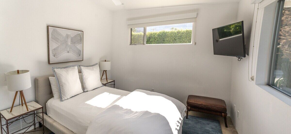 Ein Schlafzimmer mit einem Bett, Nachttischen mit Lampen, einem Fenster und einem an der Wand montierten Fernseher.