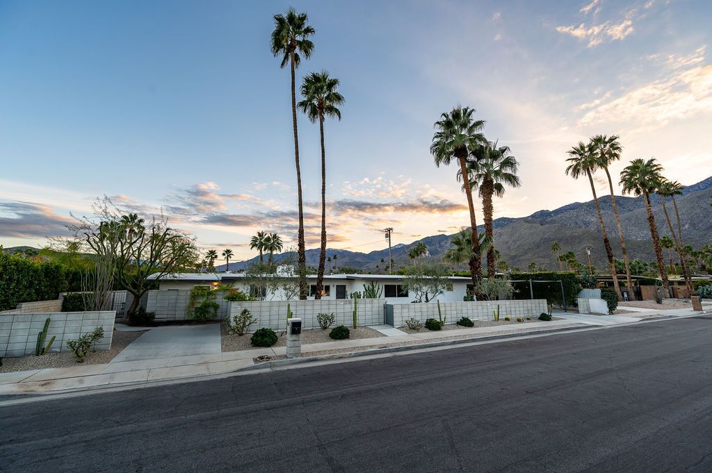 A modern house with a white exterior, surrounded by palm trees and a mountain backdrop.