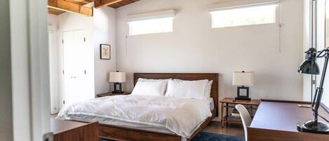 Un dormitorio con techo de madera, una cama con ropa blanca, dos mesitas de noche con lámparas, un escritorio y una silla.