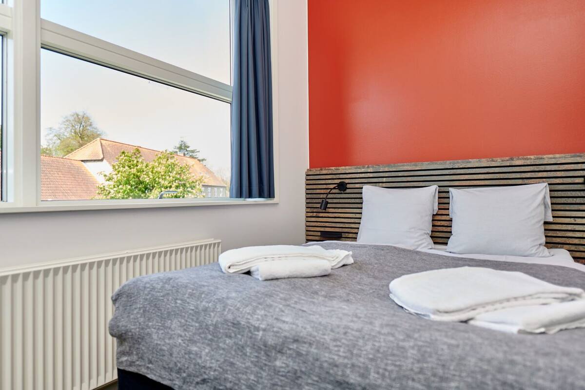 A hotel room with a bed, a window with a view of buildings, and a red wall.