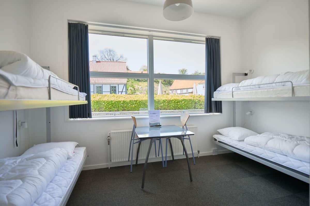 A dormitory room with bunk beds, a table, chairs, and a window with a view of buildings and greenery.