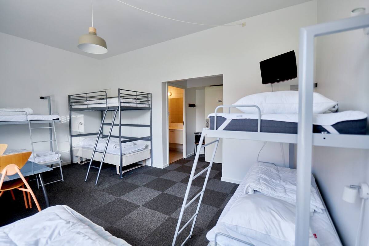 A dormitory room with bunk beds, a desk, a chair, and a TV mounted on the wall.