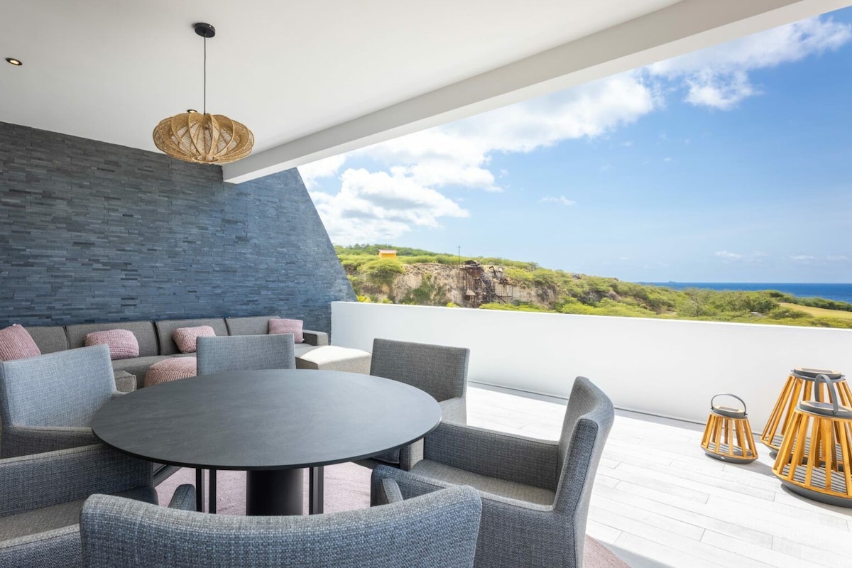 A modern outdoor seating area with a round table and chairs, a stone wall, and a view of the ocean.
