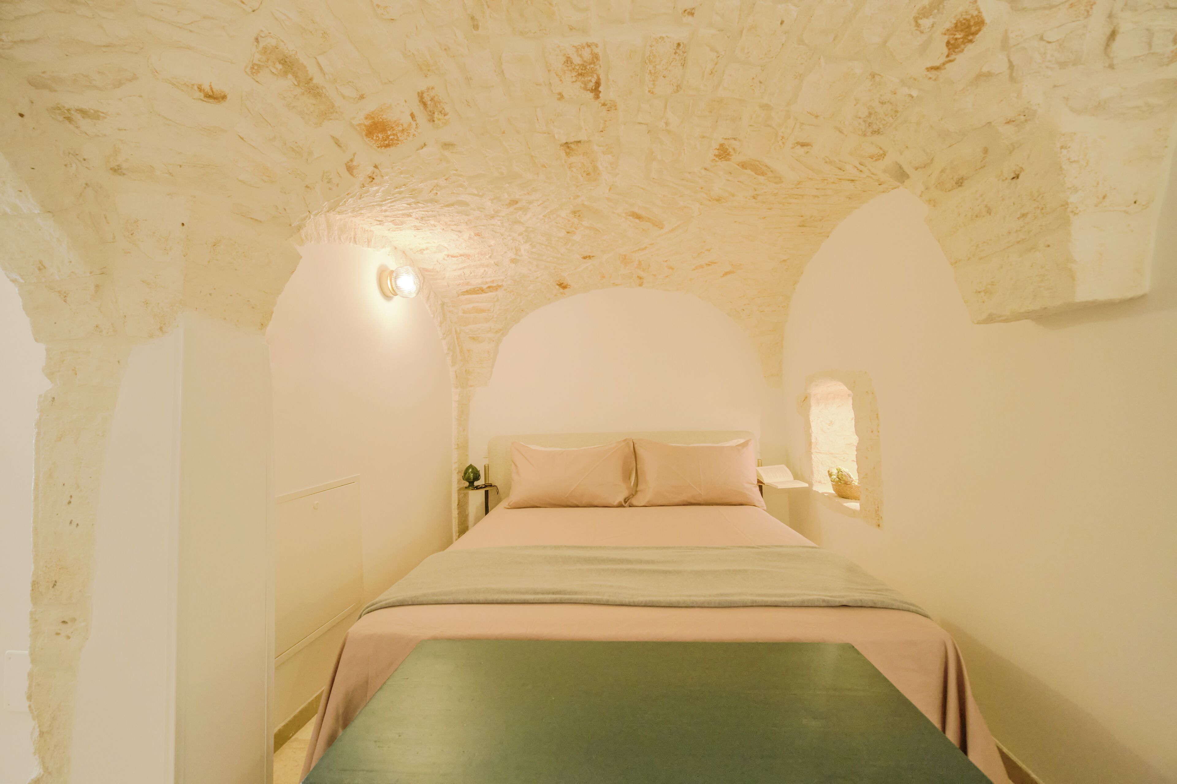 A room with a stone ceiling, a bed with a green and pink bedspread, and a small table with a lamp.