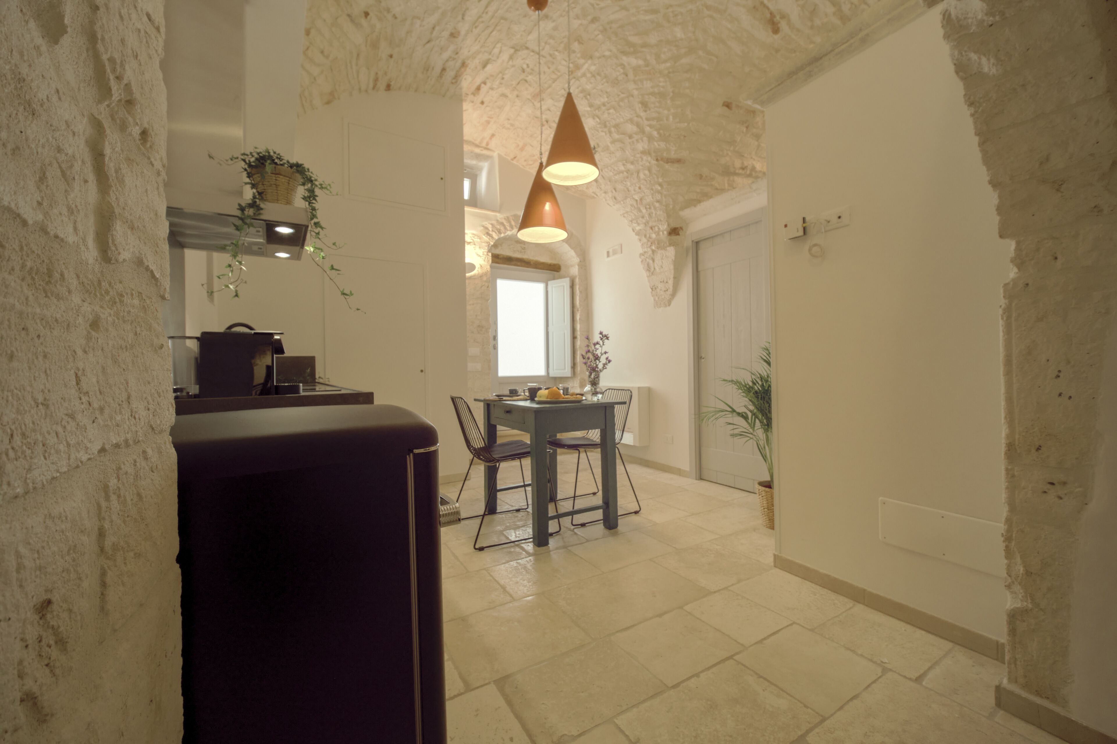 A narrow hallway with a dining area featuring a table and chairs, a hanging plant, and a stone wall.