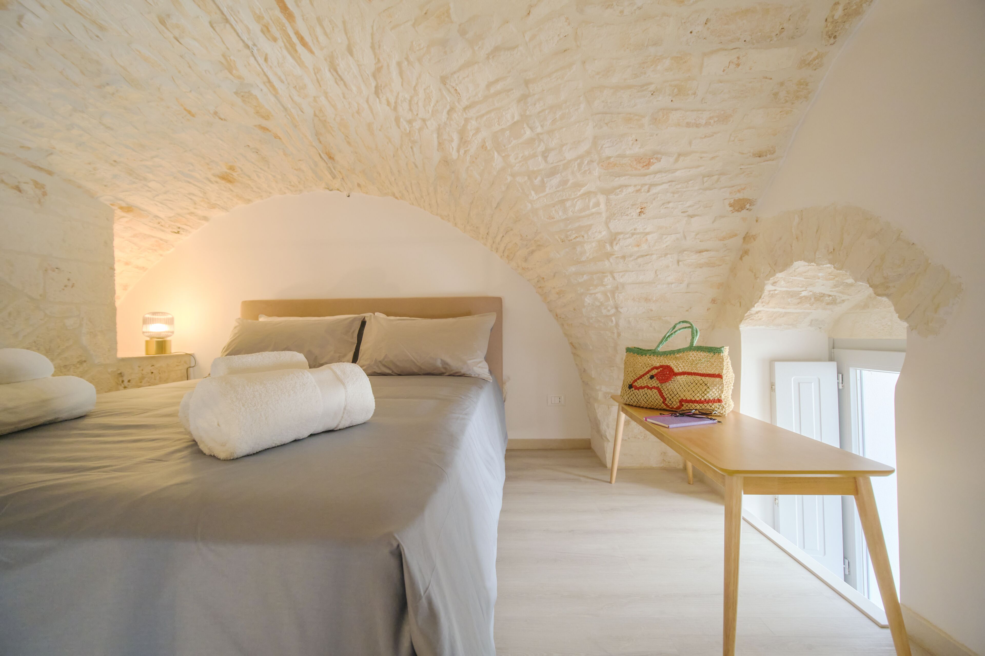 A bedroom with a stone ceiling, a bed with white linens, a bedside table with a lamp, and a window with a view.