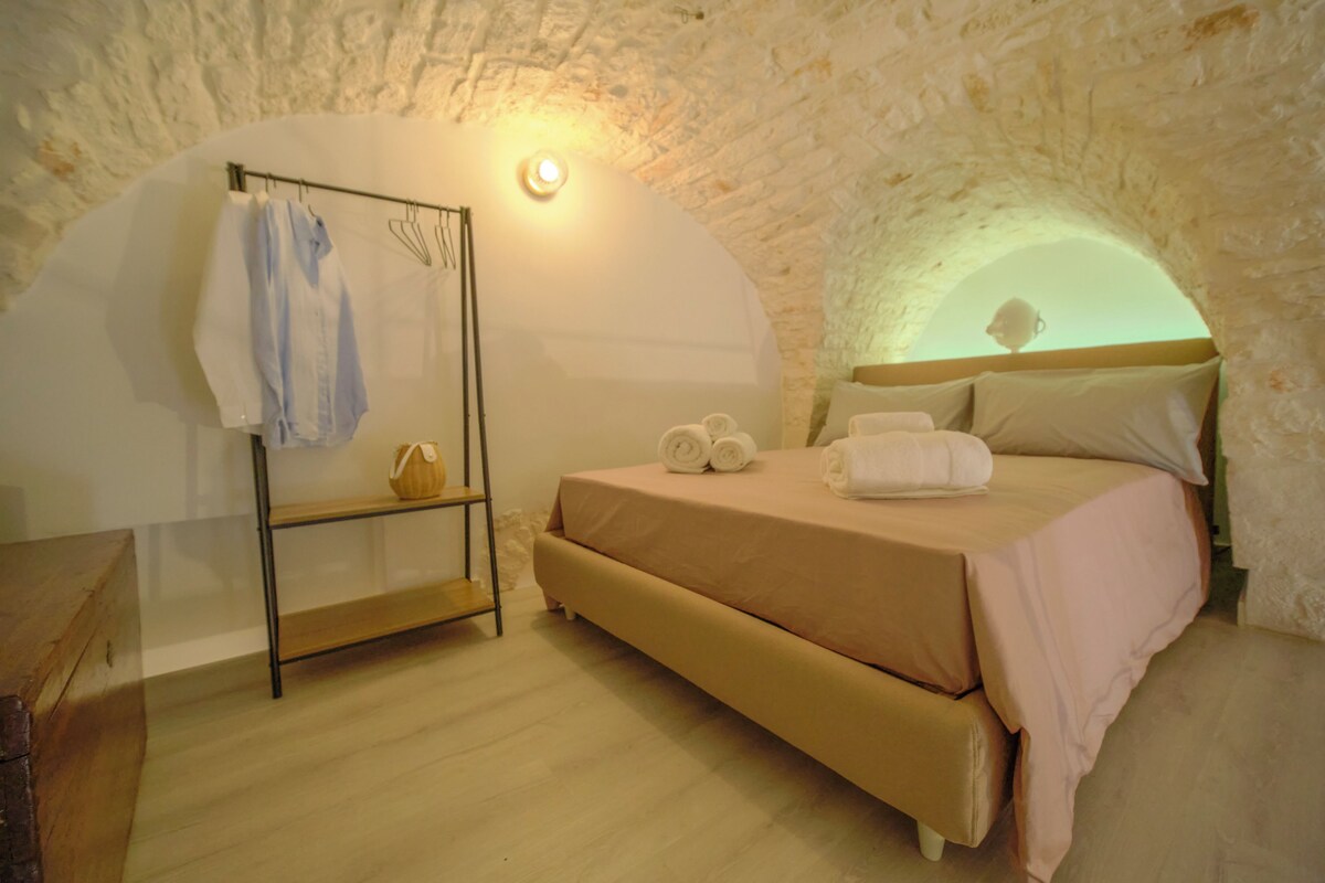 A cozy bedroom with a stone wall, a bed with a pink bedspread, a wooden shelf with towels, and a small table with a lamp.