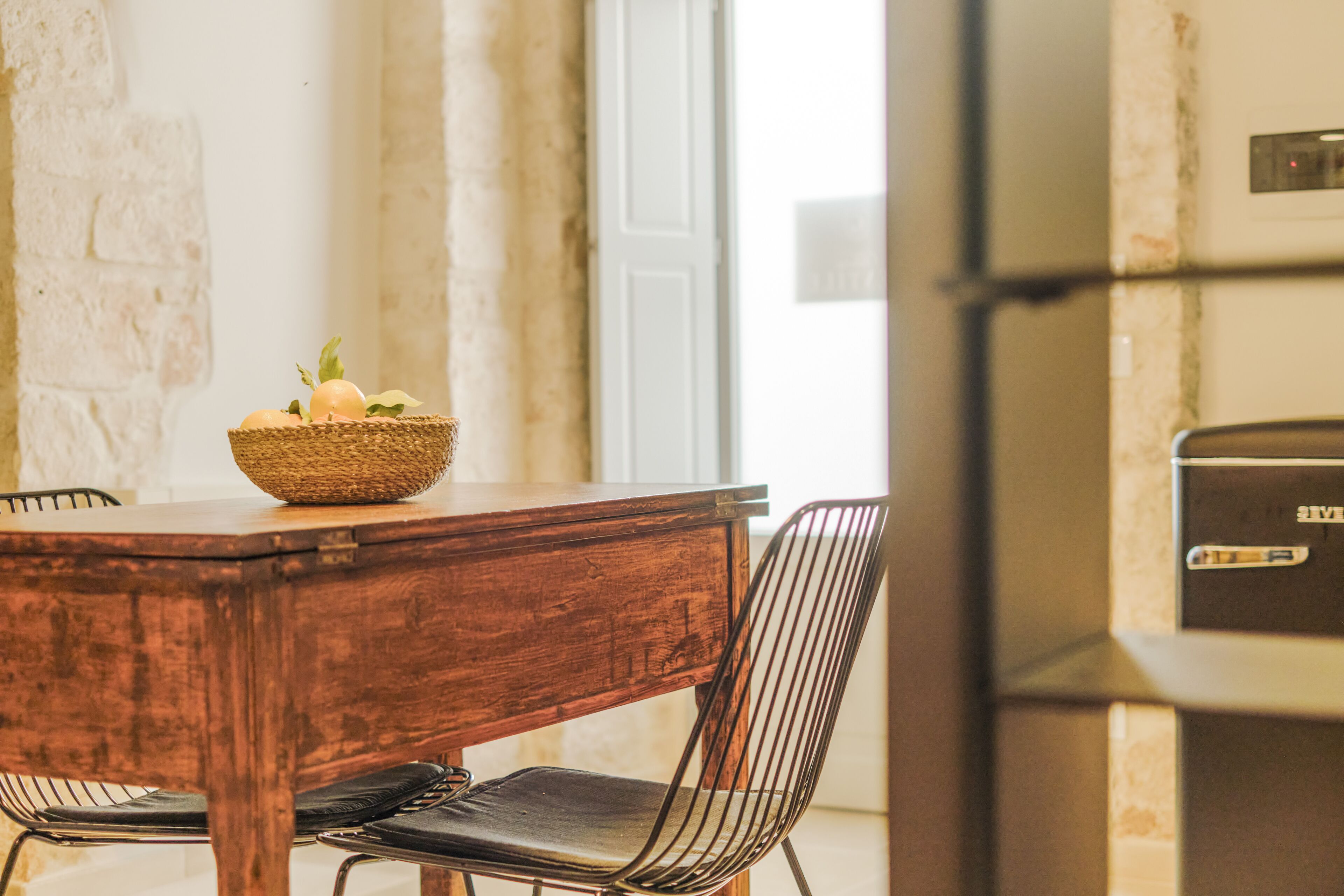 A dining area with a wooden table, a woven basket with fruits, a metal chair, and a refrigerator.