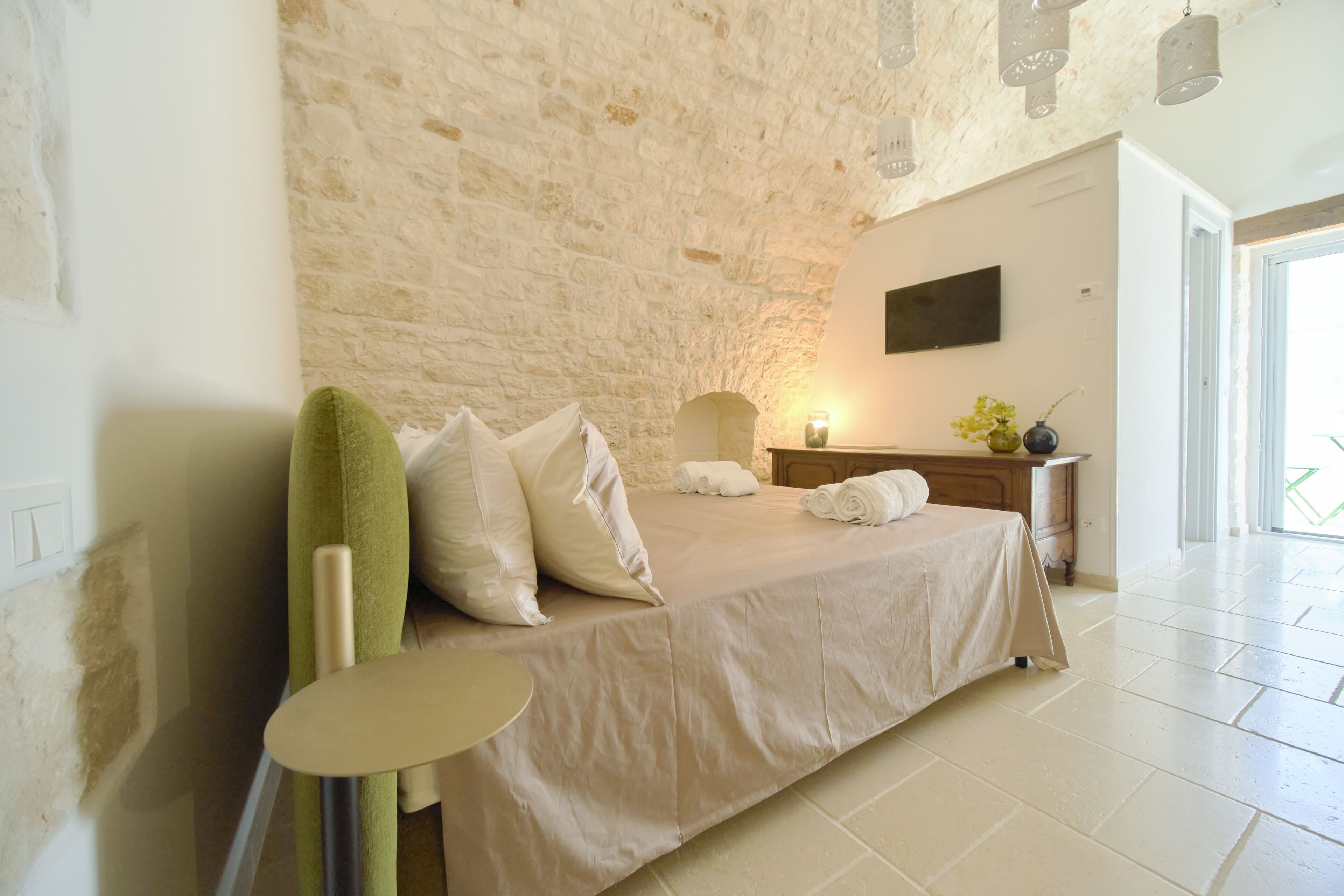 Une chambre avec un mur en pierre, un lit recouvert de draps blancs, une table de chevet avec une lampe, et une vue sur l’extérieur.