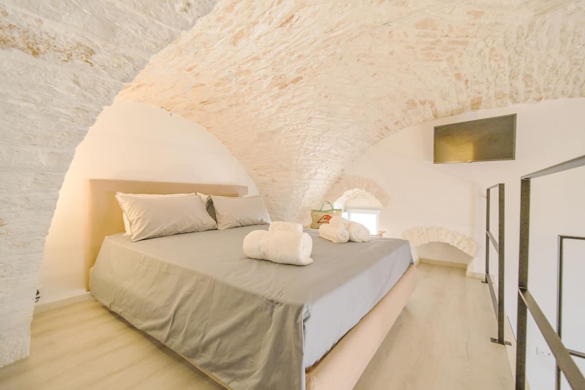 A bedroom with a stone ceiling, a bed with pillows and towels, a small table, and a wall-mounted light fixture.