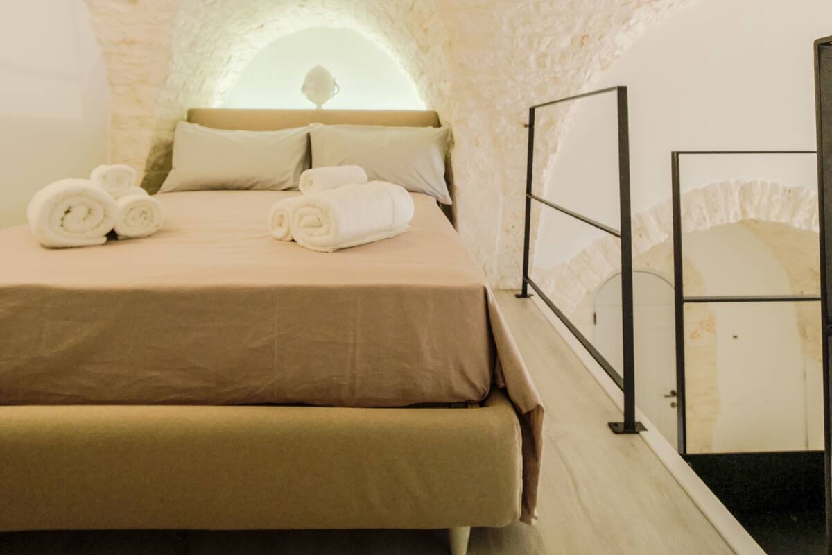 A bedroom with a stone wall, a bed with beige bedding, and rolled towels on the bed.