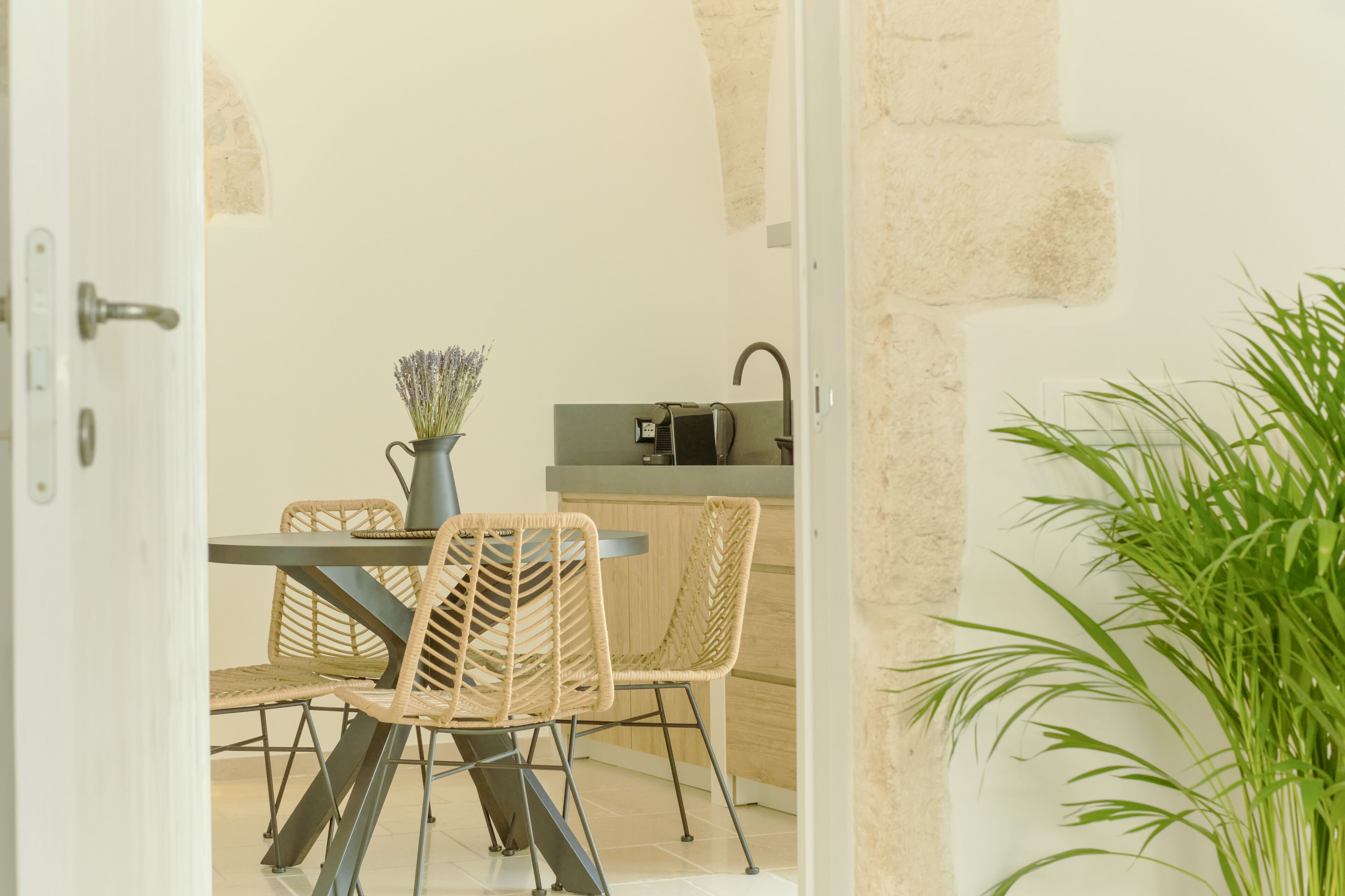 Una puerta abierta que da a un comedor con sillas de mimbre y una mesa, una planta en maceta y una cocina con un fregadero y una encimera.