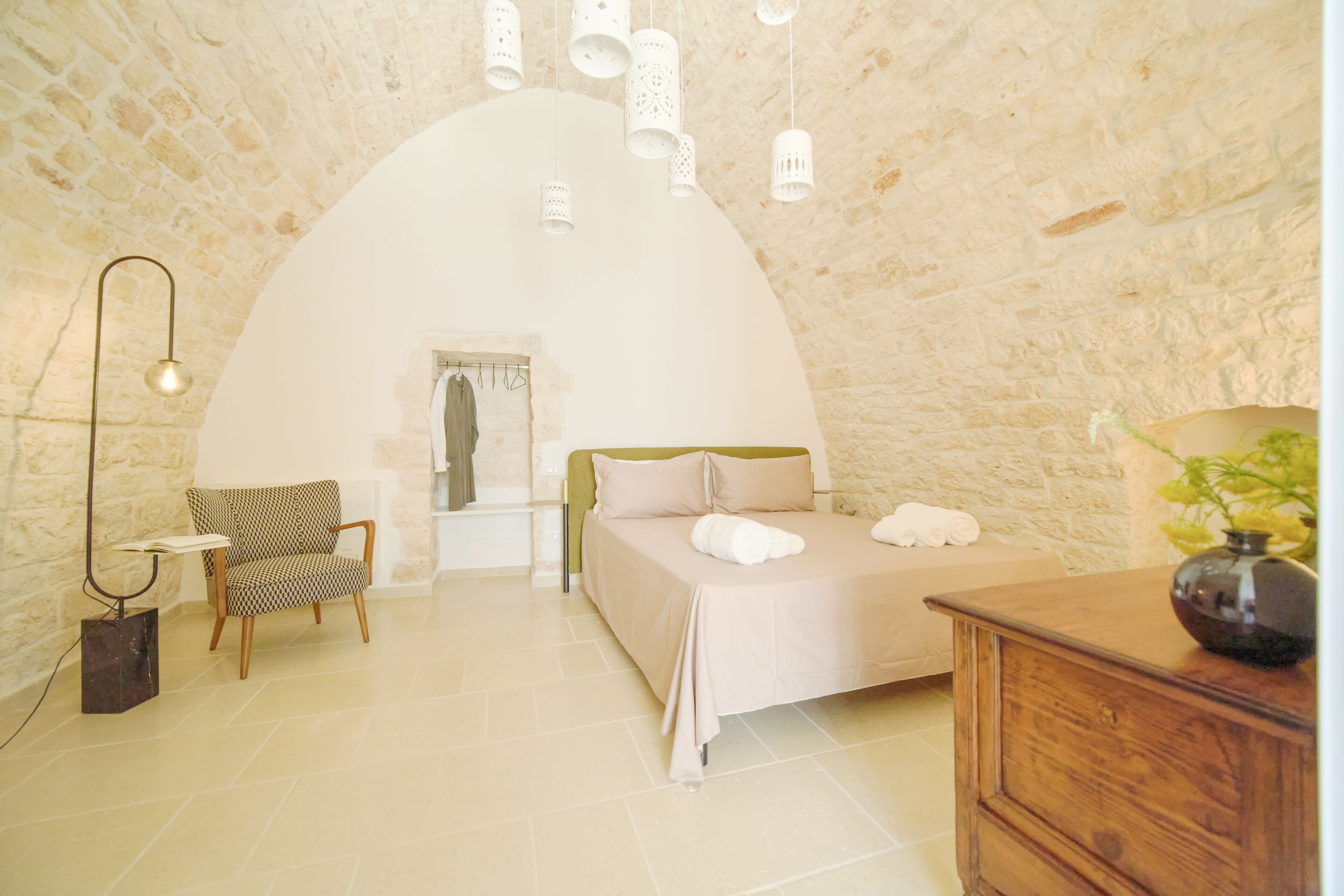A room with a high arched ceiling, a bed with white linens, a wooden chair, a wooden dresser, and a hanging light fixture.