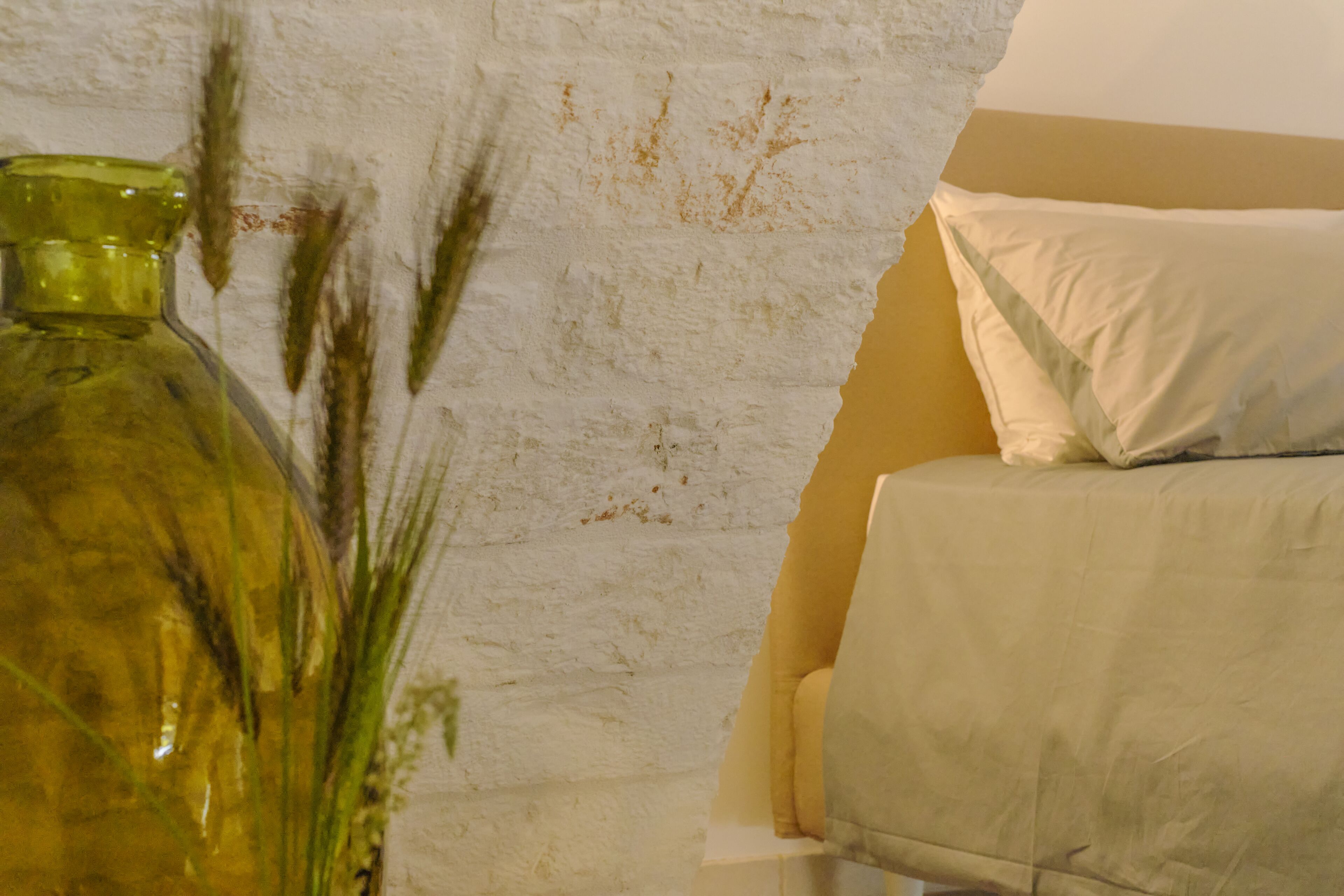A room with a white textured wall, a green vase with plants, and a bed with a white pillow.