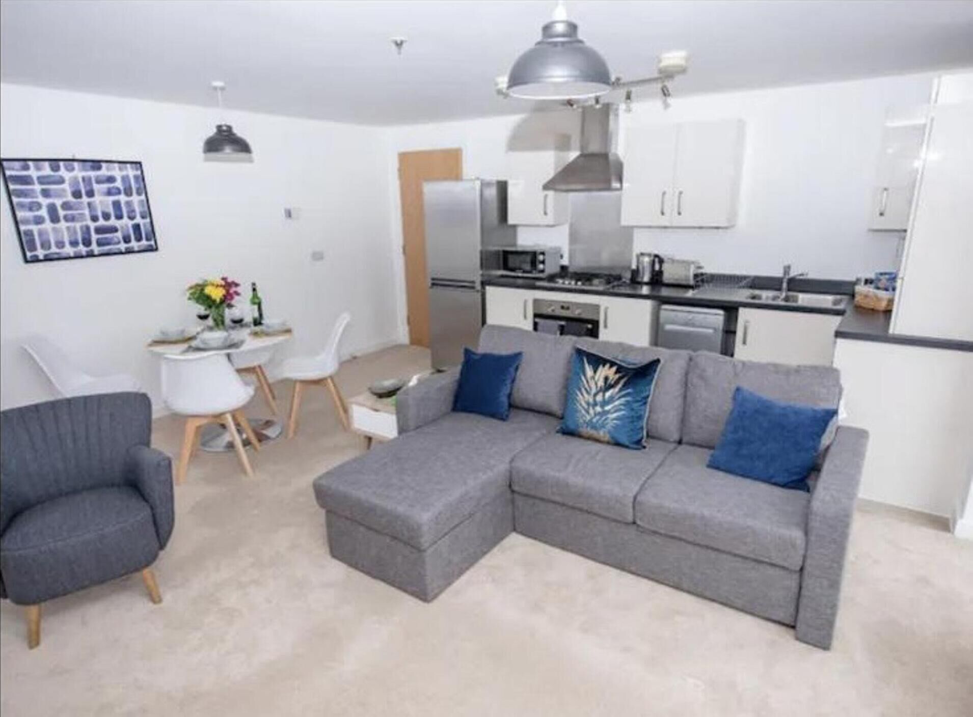 Ein modernes Wohnzimmer mit einem grauen Ecksofa, einem Essbereich mit kleinem Tisch und Stühlen sowie einer Küchenzeile mit weißen Schränken und Geräten.