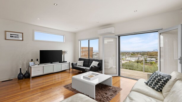 A spacious living room with a large flat-screen TV, a black leather sofa, a white coffee table, and a view of the city through large sliding glass doors.