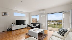 A spacious living room with a large flat-screen TV, a black leather sofa, a white coffee table, and a view of the city through large sliding glass doors.