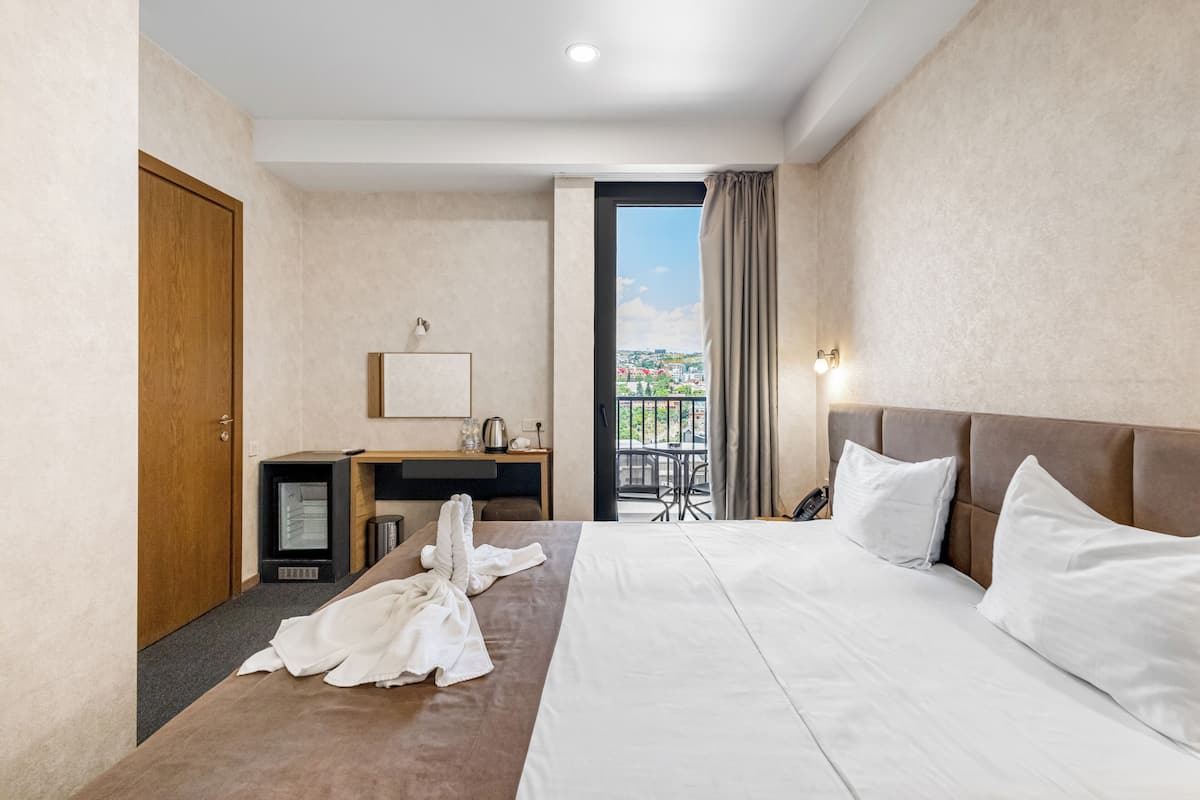 A hotel room with a bed, a desk, a chair, a small refrigerator, a window with curtains, and a view of the outside.