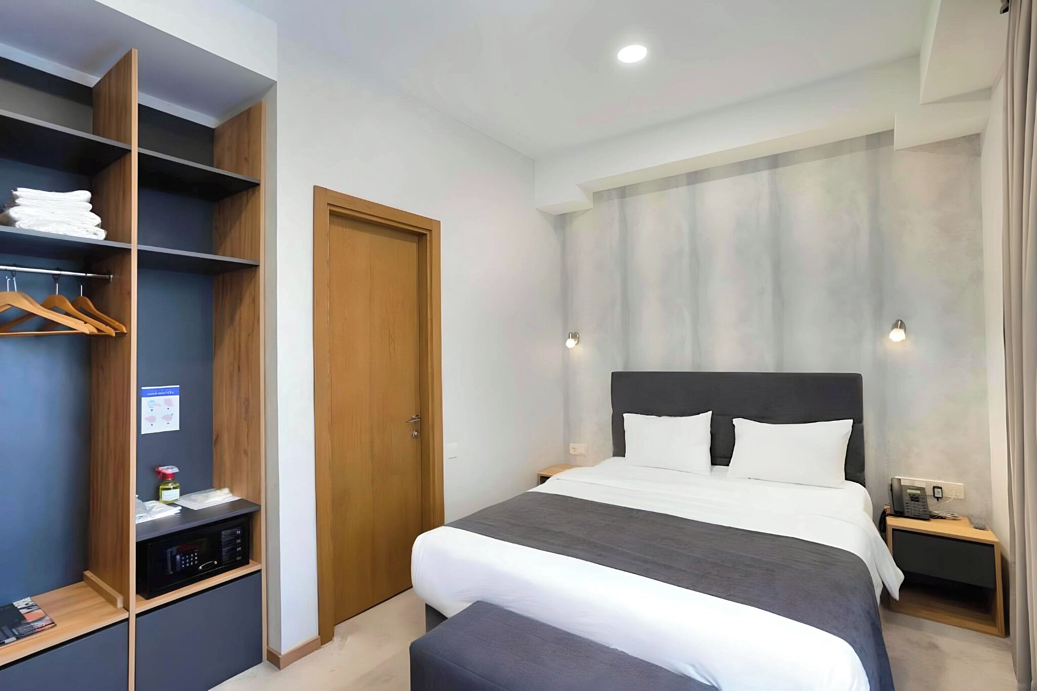 A modern hotel room with a large bed, a wardrobe with shelves, a desk with a computer, and a bedside table with a phone.