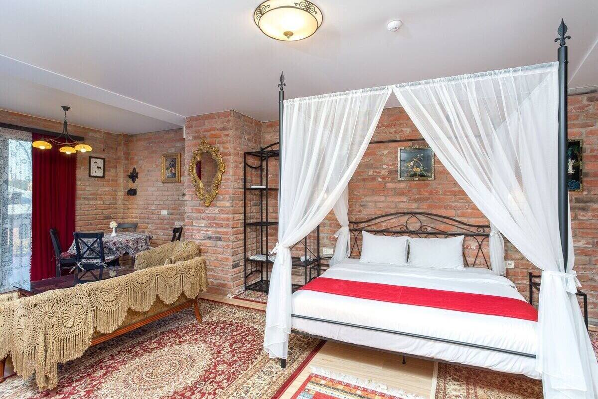 A room with a four-poster bed, a red and white bedspread, and a red curtain. There is a table with a lace tablecloth and chairs, and a patterned rug on the floor.