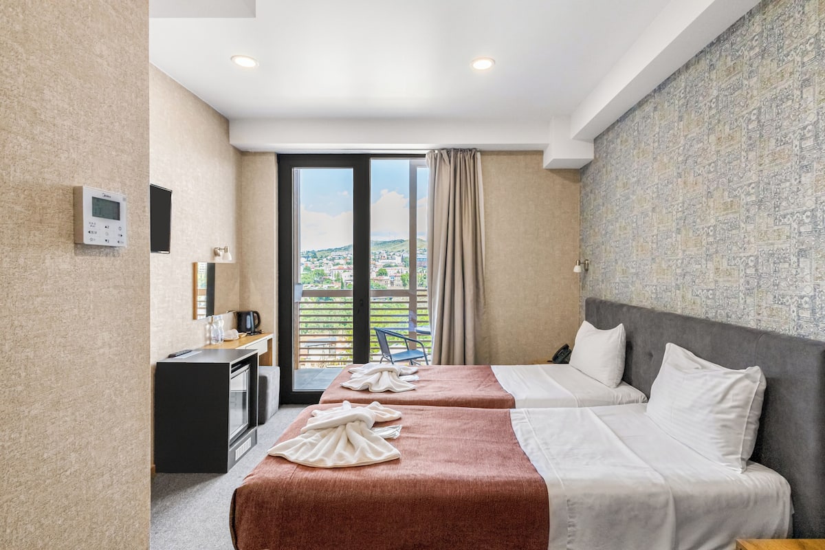 A hotel room with two beds, a desk, a coffee machine, and a view of the city.