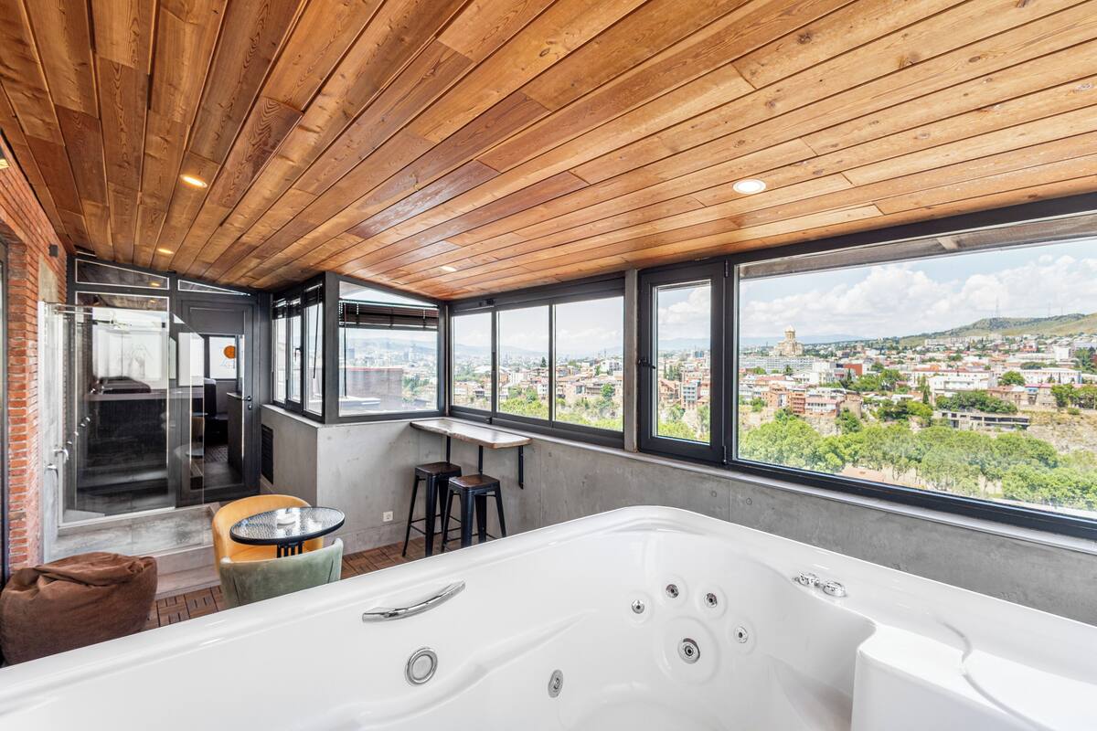 A modern bathroom with a large bathtub, wooden ceiling, and a city view through large windows.
