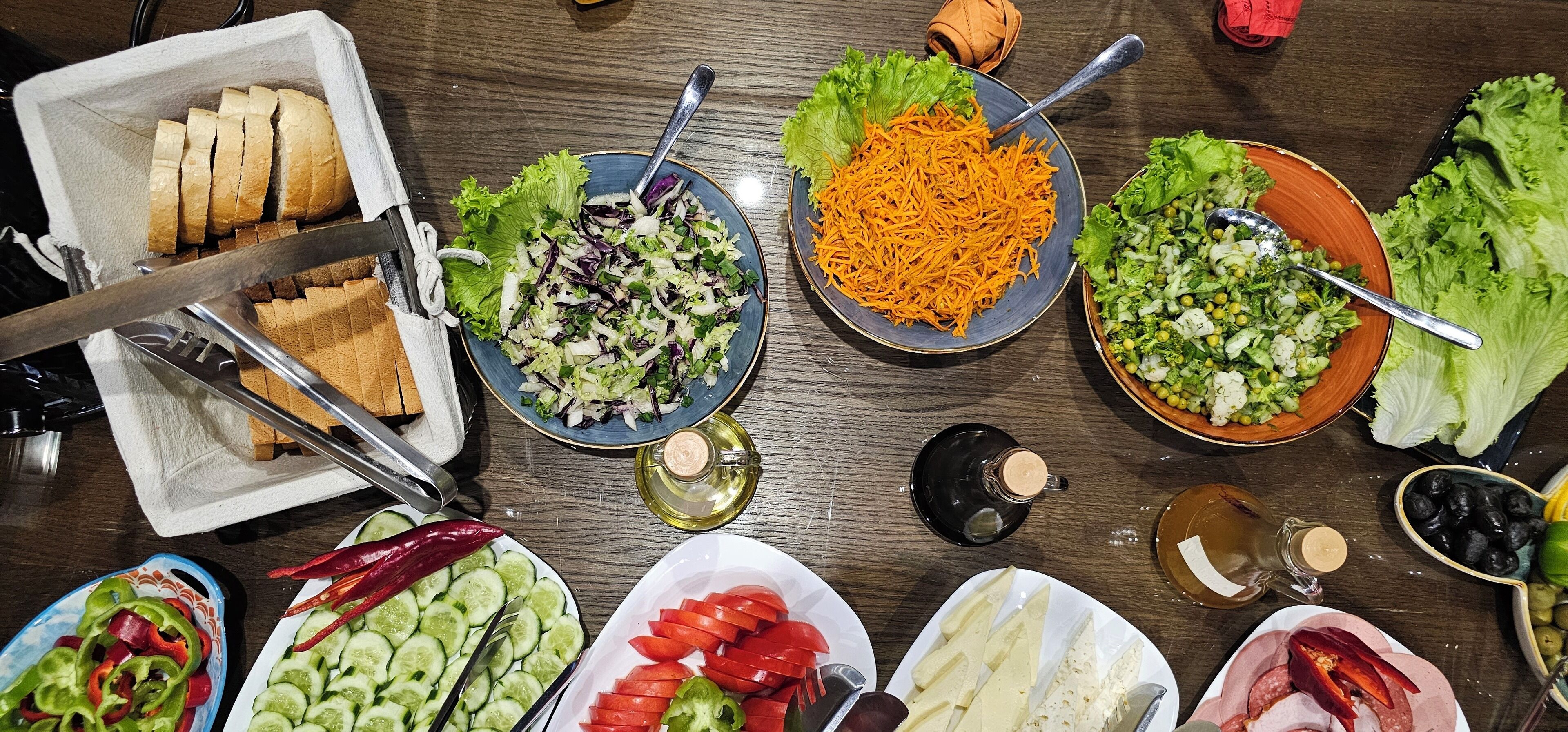 Diversos alimentos, incluindo pão fatiado, saladas e legumes, dispostos sobre uma mesa de madeira.