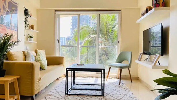 Una sala de estar con sofá, mesa de centro, una silla, televisor y un ventanal con vista a palmeras.