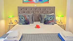 A hotel room with a tufted headboard, a colorful abstract painting above the bed, two bedside lamps, and a bed with neatly folded towels. - Okeanis Apartments & Suites Adults Only (Kefalonia)