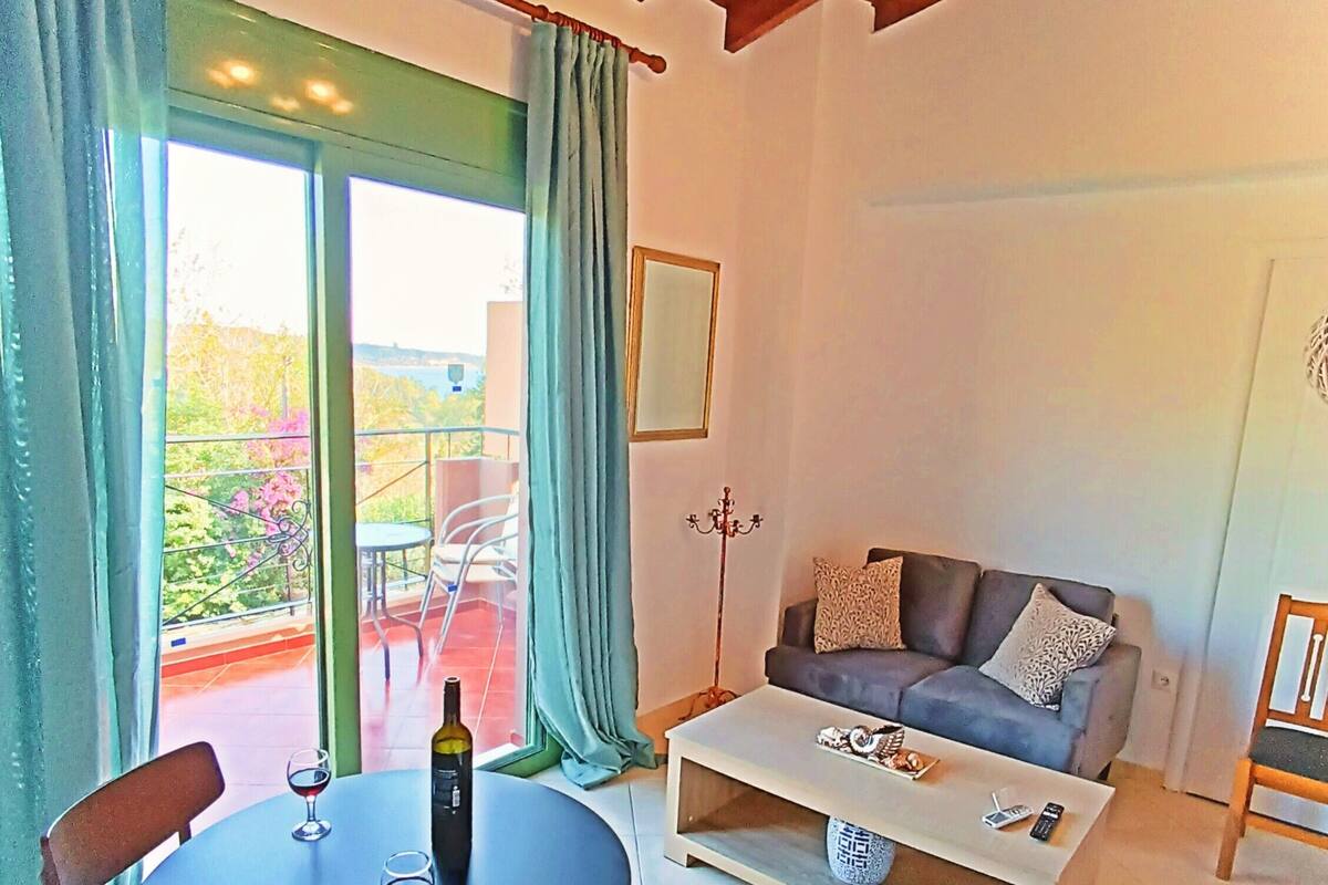 Un salon comprenant un canapé, une table basse et une table à manger avec des chaises. Il y a un balcon d’où l’on peut admirer la vue sur les arbres et un immeuble.
