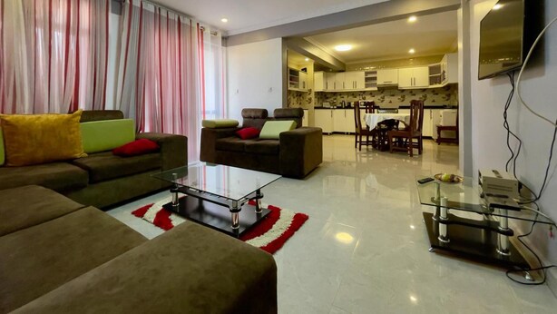 A modern living room with a sofa, ottoman, and coffee table. There is a kitchen area with cabinets and a dining table. - Charming 2-bed Apartment in Kigali (Kigali)