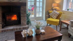 Un salon chaleureux avec une cheminée, un fauteuil jaune, une table basse avec des tasses et une théière, et une vue à travers une porte vitrée.