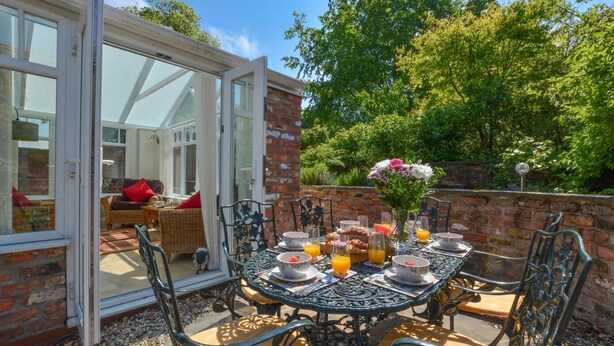 Un espace de restauration en plein air avec un mur en briques, une porte vitrée donnant sur une pièce, et une table dressée avec des assiettes, des bols et des verres remplis de jus d’orange.