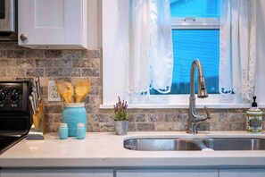 A kitchen with a stove, utensils, jars, a sink, and a window with curtains. - Blue Bungalow - Fire Pit, Yard Games & more! (Fredericksburg)