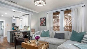 A modern living room with a sofa, coffee table, and a kitchen in the background. - Blue Bungalow - Fire Pit, Yard Games & more! (Fredericksburg)