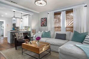 A modern living room with a sofa, coffee table, and a kitchen area in the background. - Blue Bungalow - Fire Pit, Yard Games & more! (Fredericksburg)