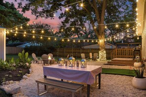 An outdoor patio with a table set for dining, string lights, and a fire pit. - Blue Bungalow - Fire Pit, Yard Games & more! (Fredericksburg)