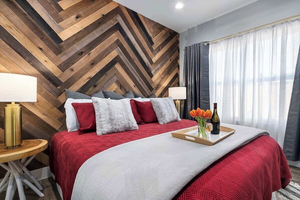 A hotel room with a herringbone wooden wall, a bed with red and white bedding, two bedside lamps, a tray with flowers and a bottle, and a window with curtains. - Strawberry Suite - Fire Pit, BBQ Grill & more! (Fredericksburg)